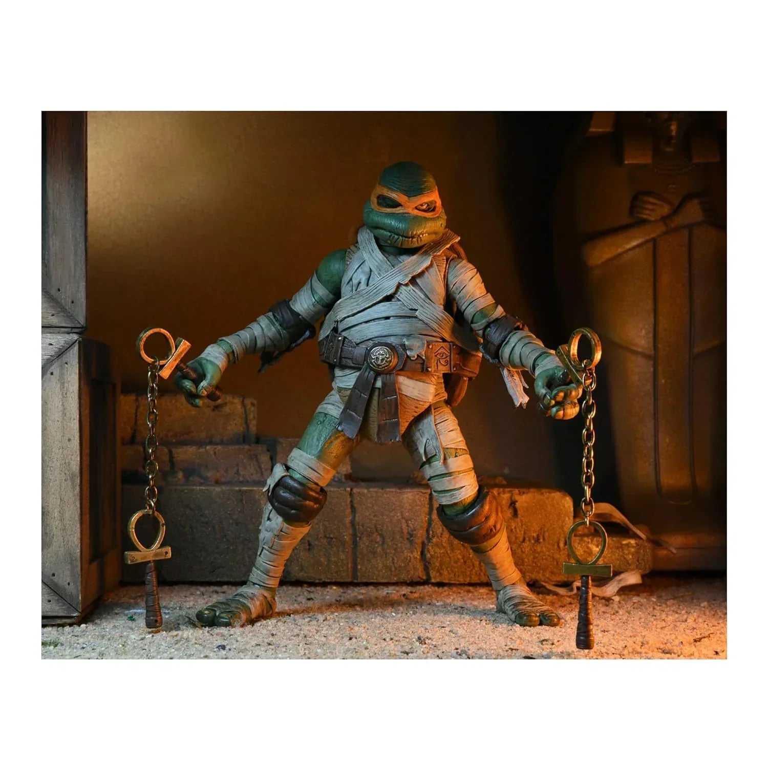 Ninja turtle action figure dressed as a mummy holding chained weapons in an ancient ruins setting.