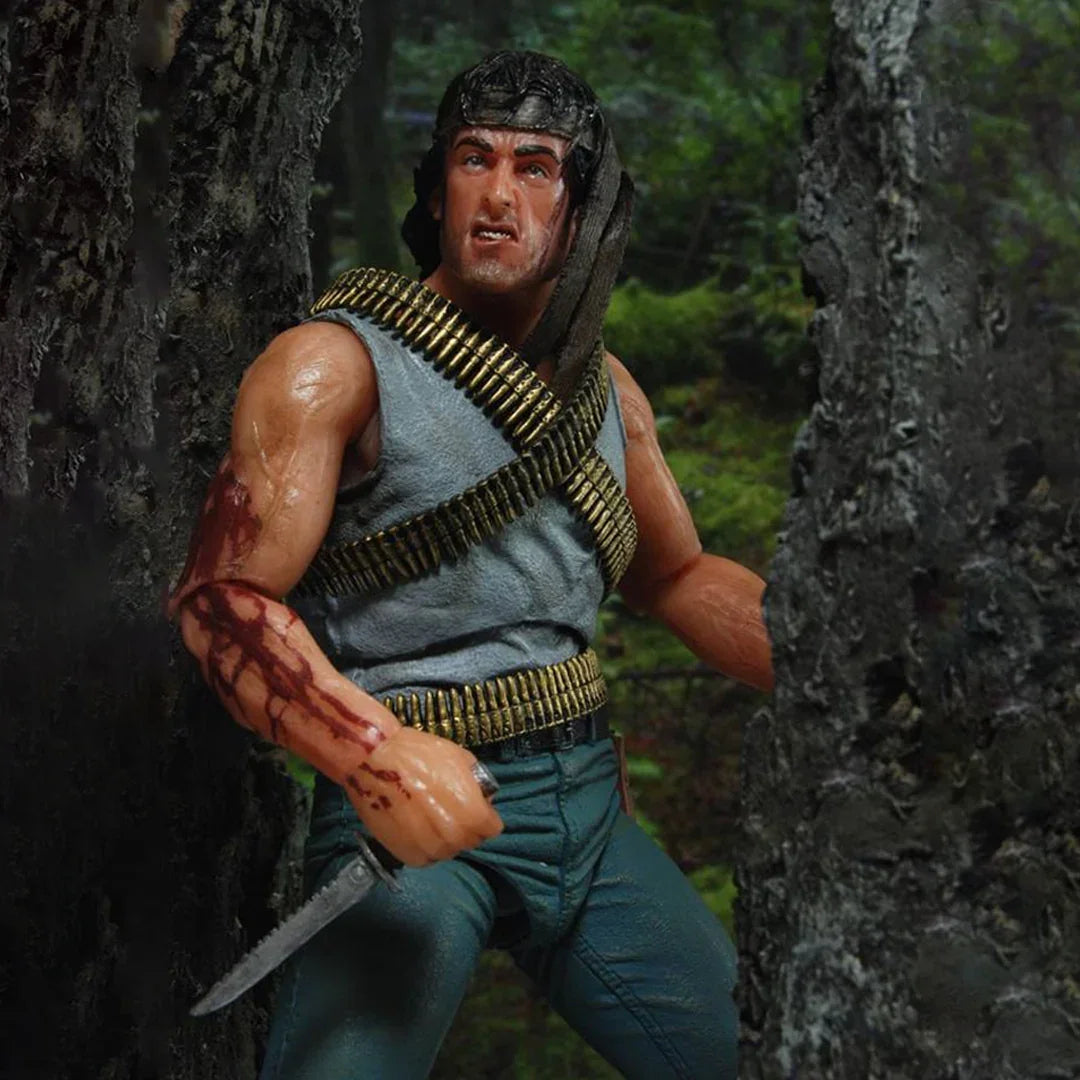 Action figure holding a knife in a forest setting, wearing ammo belts and a headband